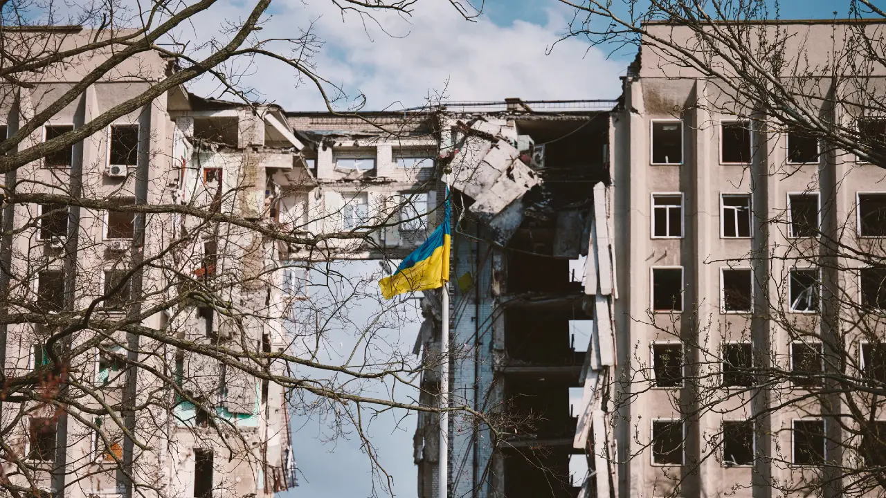 Un edificio danneggiato con un grande buco al centro e la bandiera ucraina appesa alle macerie, incorniciata da rami di alberi spogli sotto un cielo parzialmente nuvoloso.