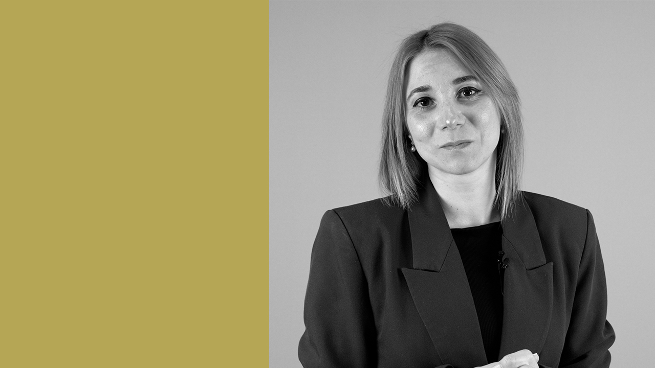 Una donna con i capelli lunghi fino alle spalle che indossa un blazer scuro si trova su uno sfondo bicolore, con una parte solida giallo senape a sinistra e una parte grigia semplice a destra. Guarda verso la telecamera con un'espressione neutra.