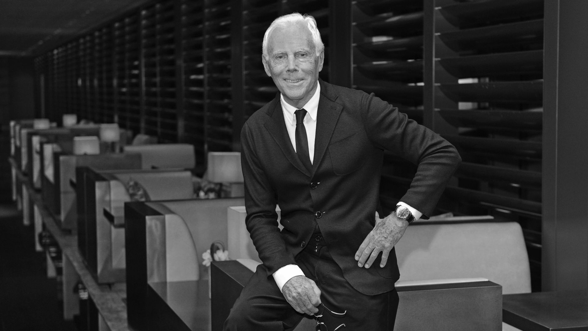 Un uomo in giacca e cravatta Armani siede con disinvoltura sul bordo di un tavolo all'interno di un ristorante moderno e di alto livello, caratterizzato da linee geometriche e illuminazione ambientale. L'immagine in bianco e nero evoca un senso di raffinatezza, un vero testamento Armani.