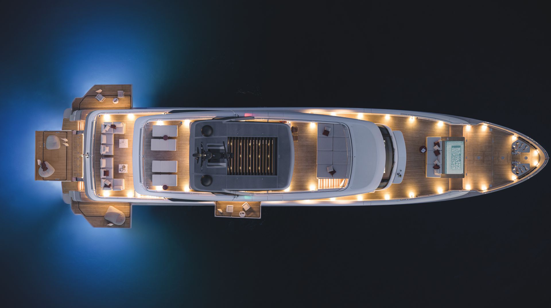 Uno yacht di lusso di Nautica Italia visto dall'alto di notte, illuminato da calde luci di coperta e luci subacquee blu, con eleganti aree di seduta e una piscina visibile sullo spazioso ponte in legno.