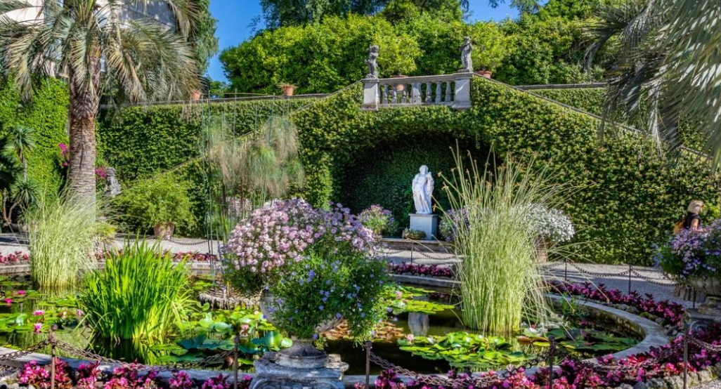 Giardino lussureggiante con stagno circolare pieno di ninfee, circondato da fiori rosa ed erbe alte. Una statua bianca di Borromeo si trova davanti a un muro verde ricoperto di edera, sotto un cielo blu brillante.