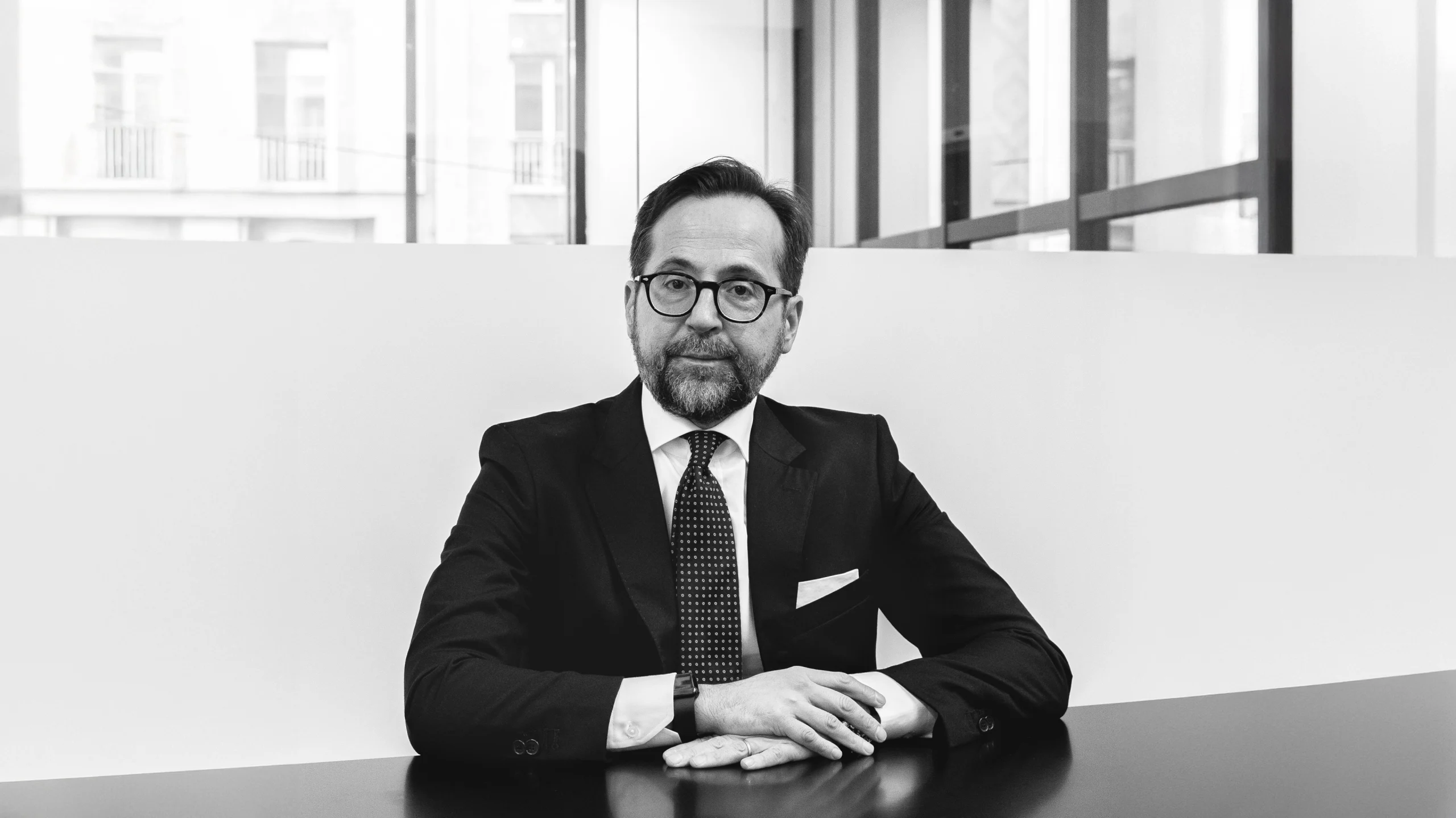 Un uomo in giacca e cravatta siede a un tavolo con le mani giunte, guardando la telecamera. Ha gli occhiali, la barba e si trova in un ufficio moderno con grandi finestre. L'immagine in bianco e nero riflette la professionalità di Kruso Kapital.