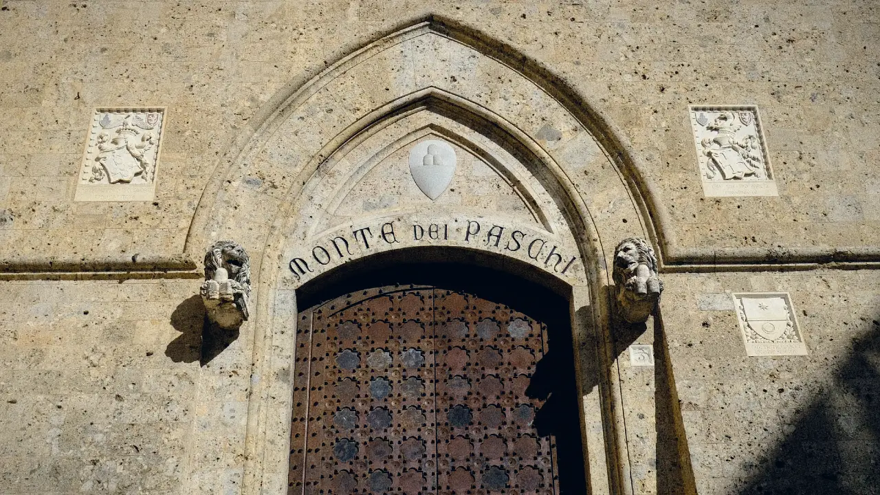 Un arco in pietra con l'iscrizione "Monte dei Paschi" sopra un grande portone in legno, affiancato da due teste di animali intagliate e da targhe decorative in pietra su entrambi i lati del muro danneggiato dalle intemperie.