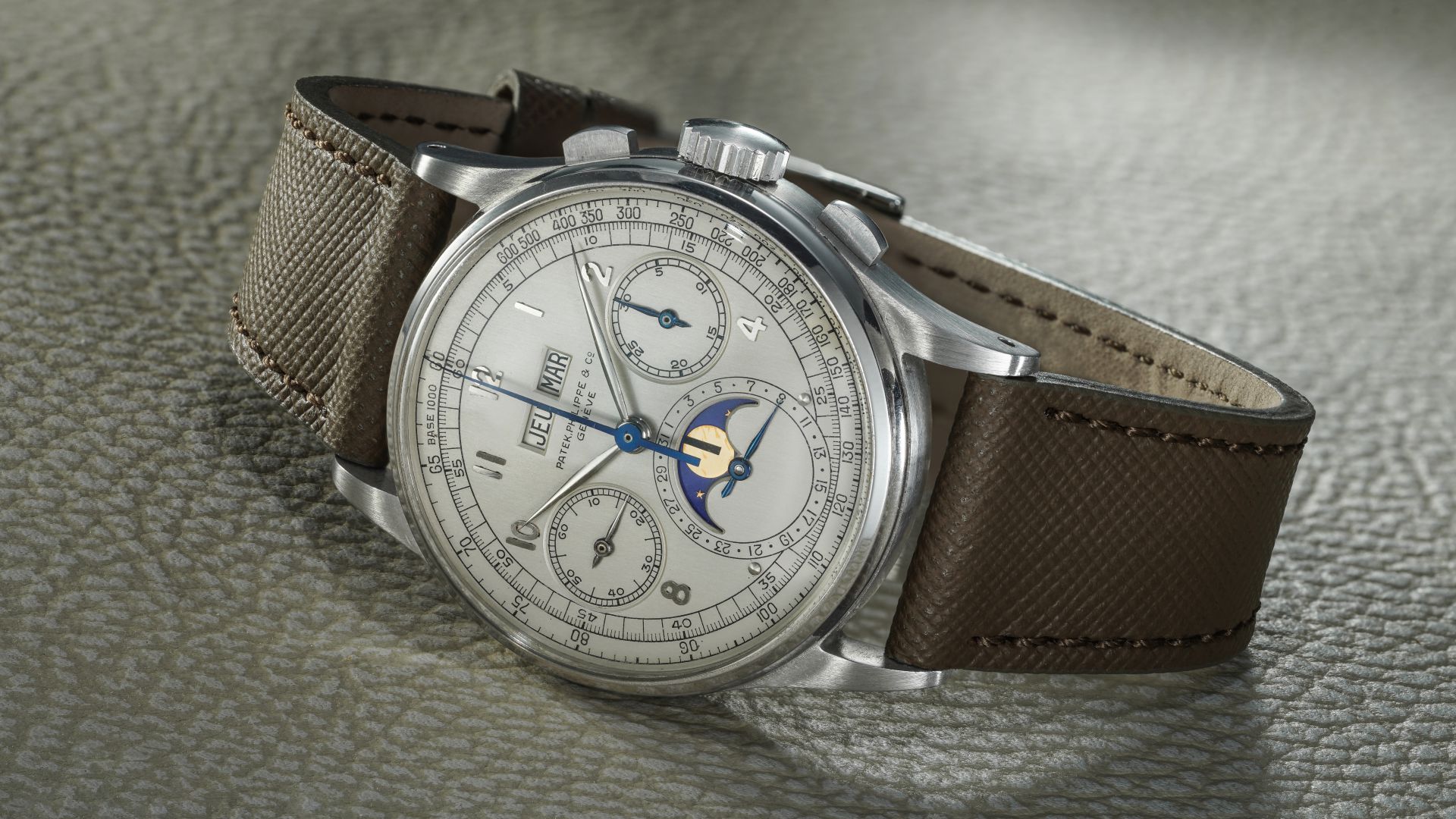 Primo piano di un lussuoso orologio da polso Patek Philippe 1518 con quadrante argentato, tre contatori, indicazione delle fasi lunari e cinturino in pelle testurizzata marrone, appoggiato su una superficie testurizzata grigio chiaro: perfetto per qualsiasi asta di alto profilo.