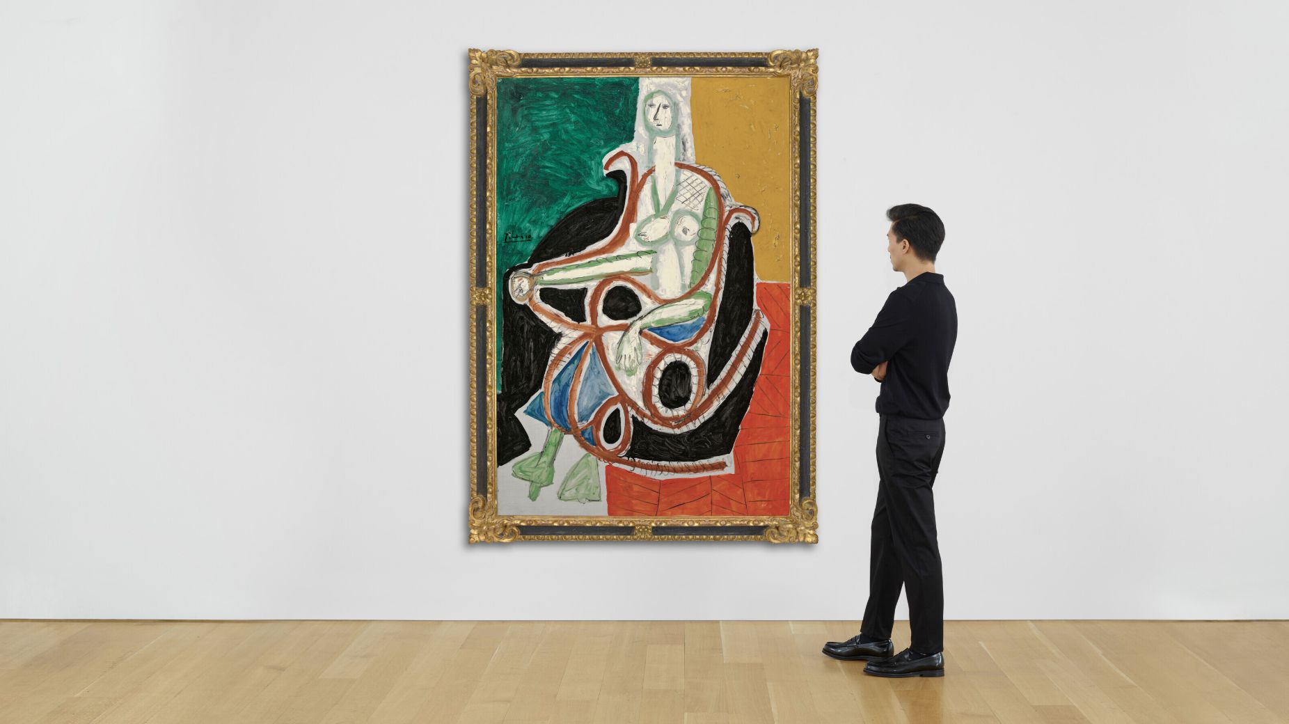 Un uomo in piedi, con le braccia incrociate, guarda un grande dipinto astratto in una cornice dorata su una parete della galleria. L'opera, che ricorda lo stile di Picasso, presenta una figura seduta con forme geometriche e colori vivaci.