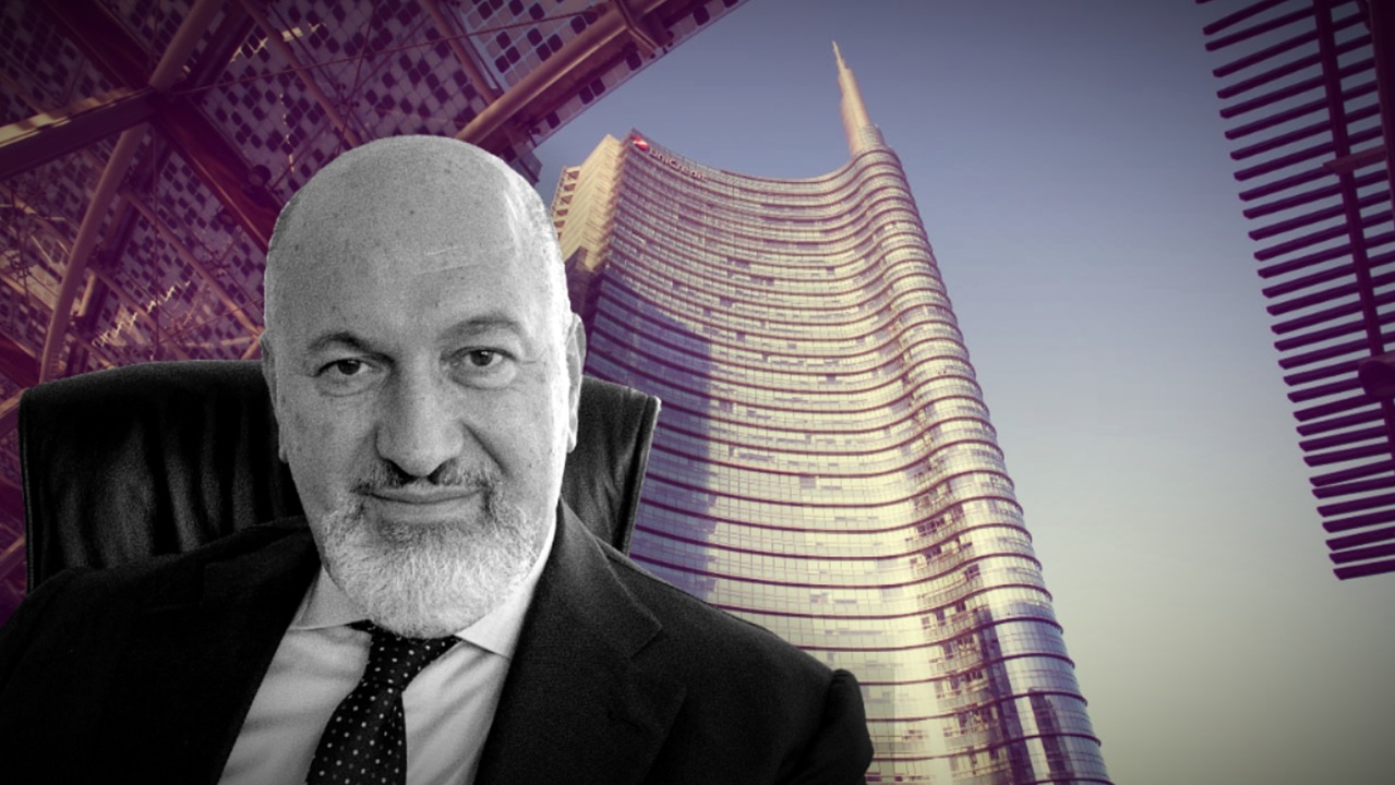 Un uomo in giacca e cravatta è seduto in primo piano, mentre sullo sfondo un moderno grattacielo di vetro curvo si staglia nel cielo. L'immagine ha un'atmosfera professionale e urbana.