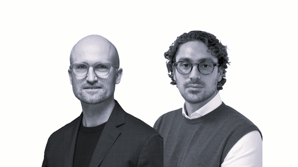 Two men wearing glasses pose in business attire against a plain white background. The man on the left is bald and wears a checked jacket, while the man on the right has wavy hair and wears a sweater over a collared shirt.