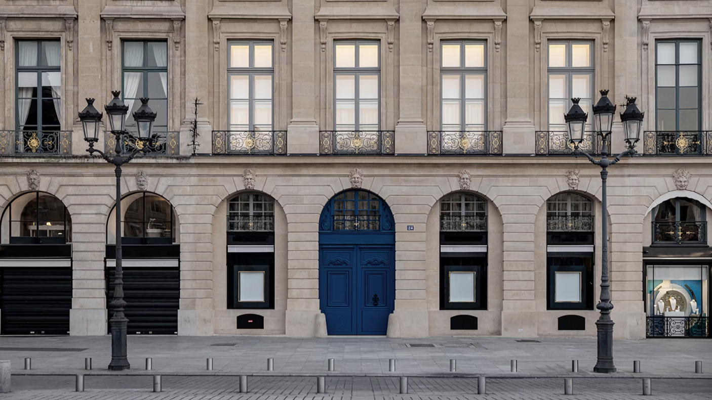 Un edificio simmetrico in pietra beige con finestre ad arco e una doppia porta centrale blu; lampioni neri e ringhiere metalliche costeggiano il marciapiede vuoto di fronte.
