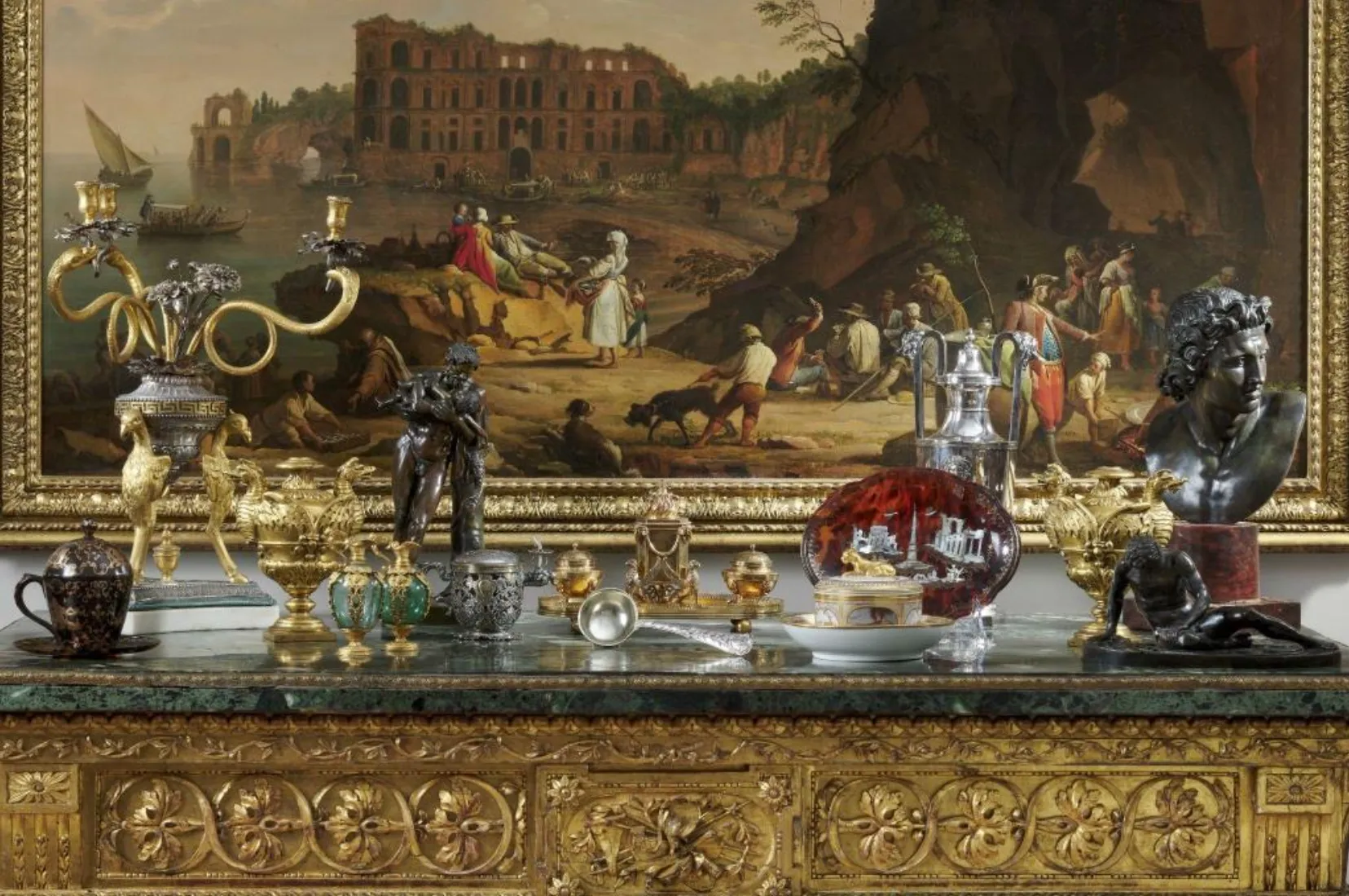 Un tavolo d'oro ornato con candelabri, argenteria e oggetti decorativi elaborati si trova sotto un dipinto classico dettagliato di persone vicino ad antiche rovine. La scena è ricca di oro, marmo e intricata maestria.