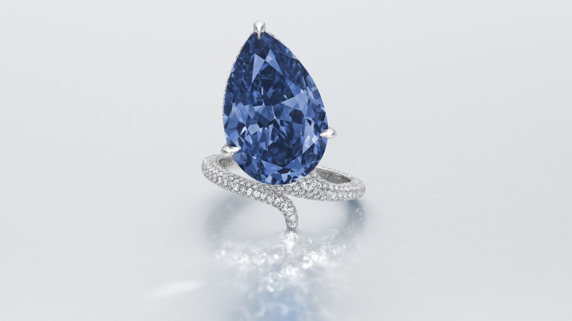 Anello con gemma blu diamantata a forma di pera, con fascia d'argento ritorta e incrostata di piccoli diamanti bianchi, su una superficie leggermente riflettente.