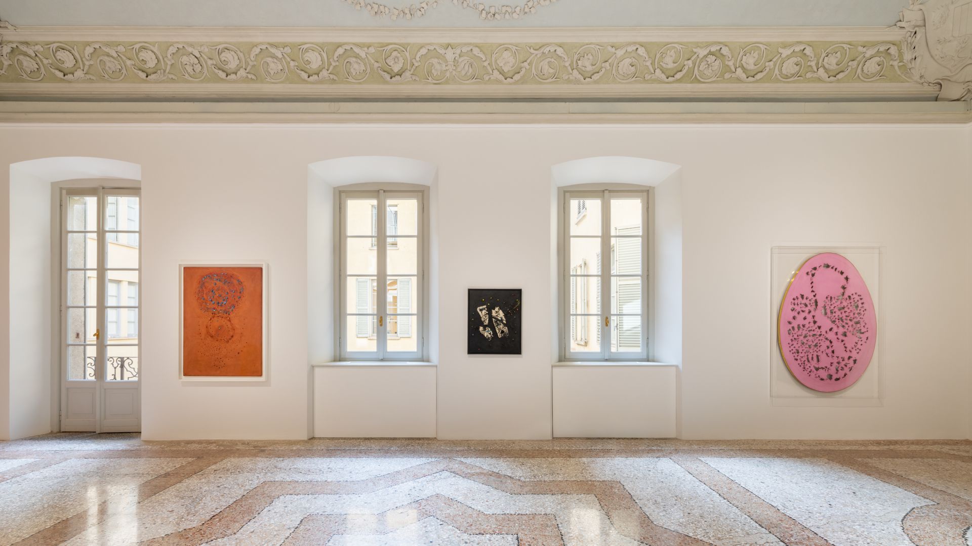 La luminosa sala della galleria di Thaddeus Ropac Milano è caratterizzata da tre alte finestre e da colorate opere d'arte astratta - un pezzo arancione, un piccolo pezzo scuro e un grande pezzo ovale rosa - appese a una parete bianca sotto un soffitto ornato.