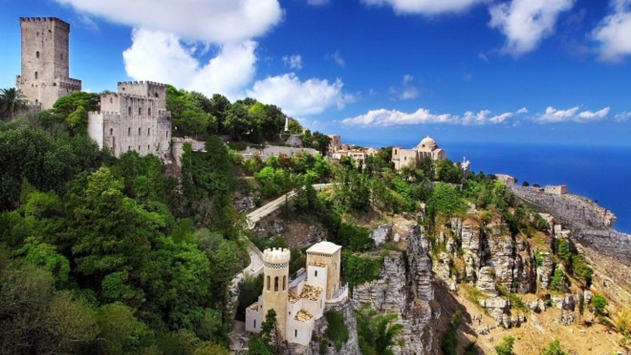 Una vista panoramica di un castello medievale in pietra vicino a Trapani, circondato da alberi verdeggianti, con scogliere drammatiche e il mare blu sullo sfondo sotto un cielo parzialmente nuvoloso.