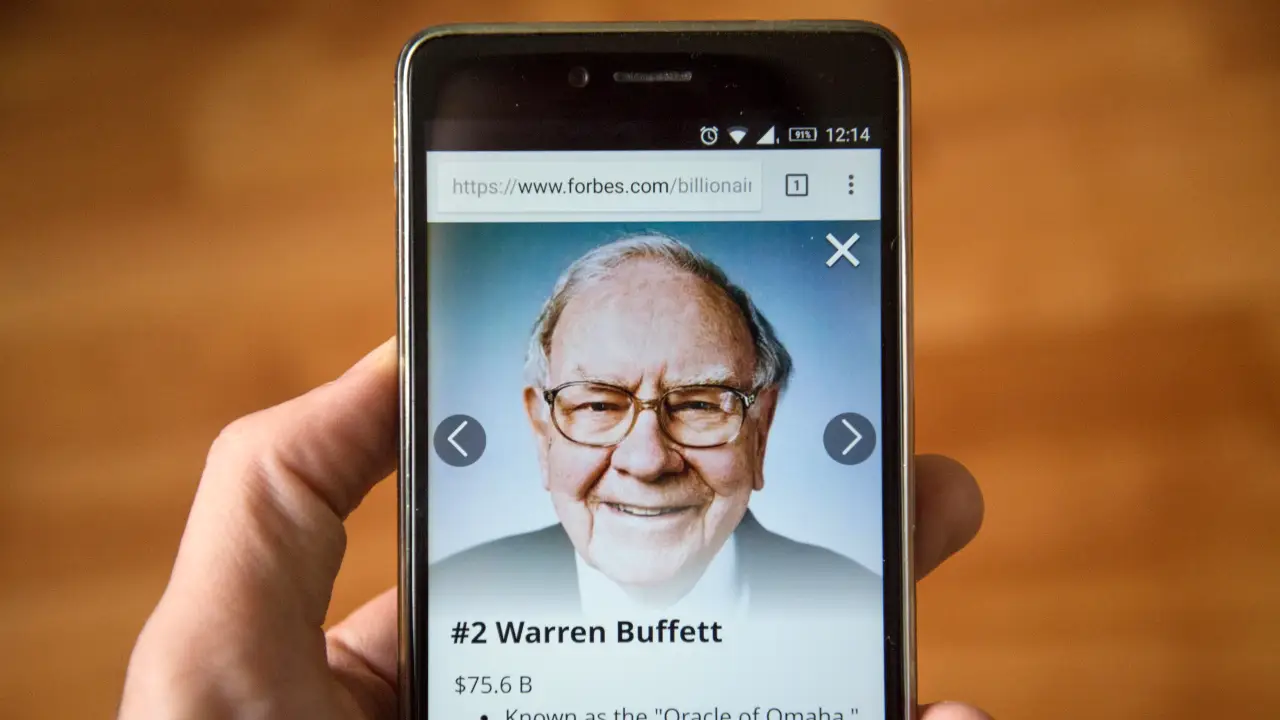 Una mano che tiene uno smartphone che mostra una foto di Warren Buffett con il testo "#2 Warren Buffett" e il suo patrimonio netto, su una pagina del sito web di Forbes.