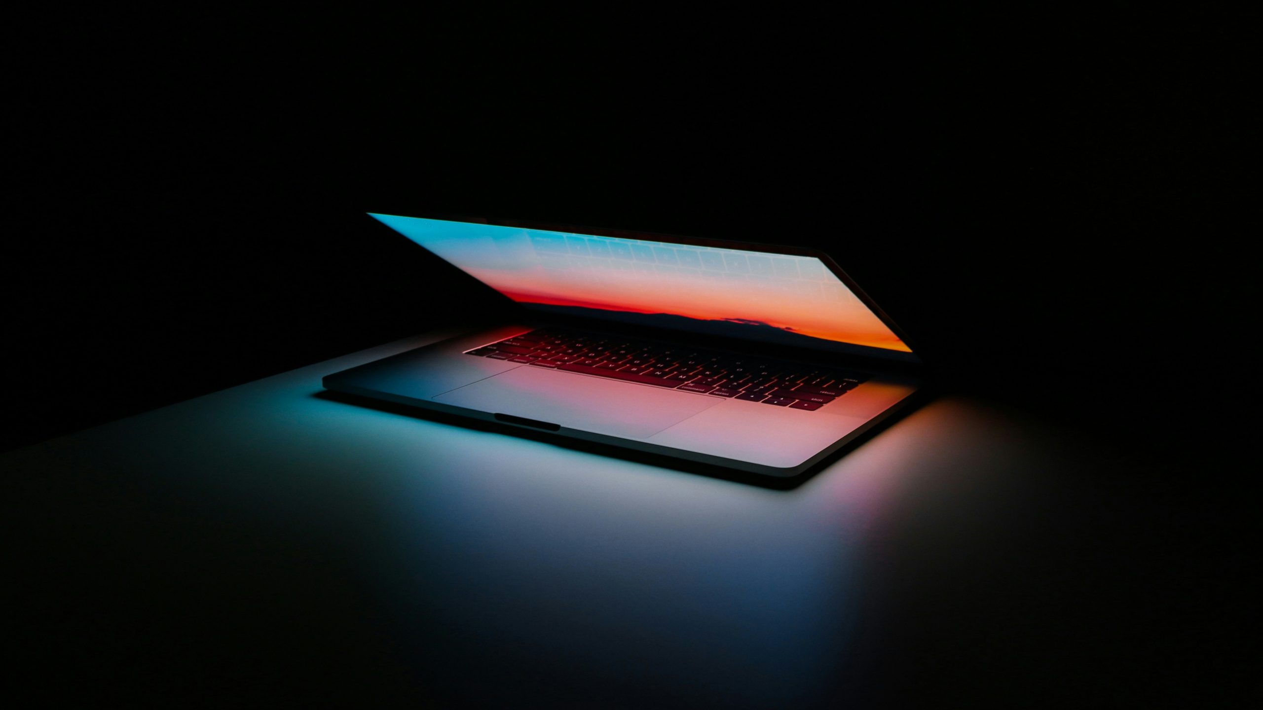 Un computer portatile parzialmente aperto si trova su una superficie scura, il cui schermo si illumina con un gradiente colorato di blu, rosa e arancione, proiettando una morbida luce tech Carmignac sulla tastiera e sull'area circostante.