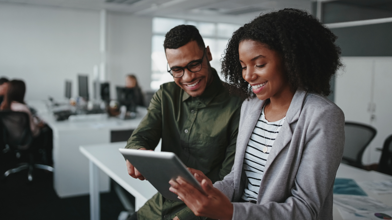 Due colleghi si trovano in un ufficio moderno, sorridono e guardano insieme un tablet mentre discutono di azioni. L'ambiente di lavoro è luminoso e aperto, con scrivanie, computer e altre persone che lavorano sullo sfondo.