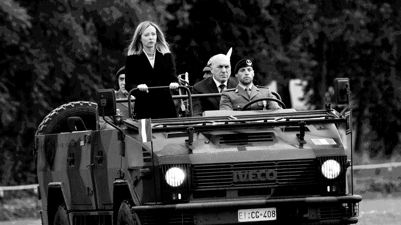 Una foto in bianco e nero mostra una donna e tre uomini in uniforme militare a bordo di un veicolo militare Iveco, con alberi sfocati sullo sfondo. La donna è in piedi davanti, con un'aria seria.