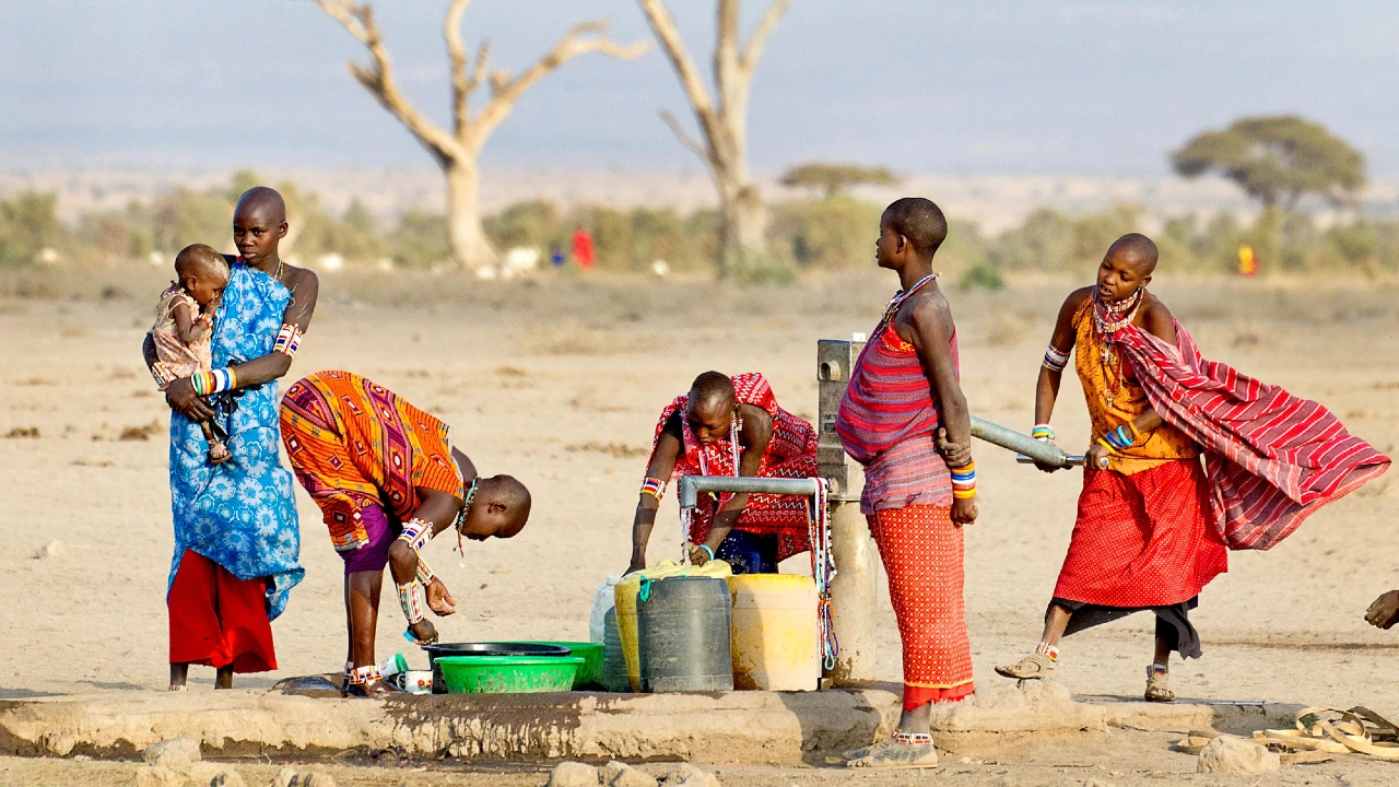 Quattro donne in abiti tradizionali colorati raccolgono acqua da una pompa in un paesaggio arido e aperto; una tiene in braccio un bambino, mentre altre riempiono dei contenitori. Si intravedono alberi lontani e uno sfondo nebuloso.