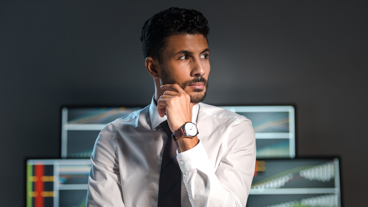 Un uomo in camicia bianca e cravatta se ne sta pensieroso con la mano sul mento, guardando di lato. Sullo sfondo, i monitor di un computer mostrano grafici e tabelle che tracciano l'andamento dei mercati.