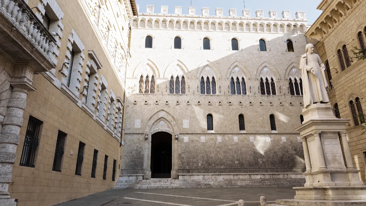 Un edificio storico in pietra con finestre ad arco e intagli dettagliati si trova in una piazza illuminata dal sole. Una statua di marmo bianco su un piedistallo è visibile sul lato destro dell'immagine.