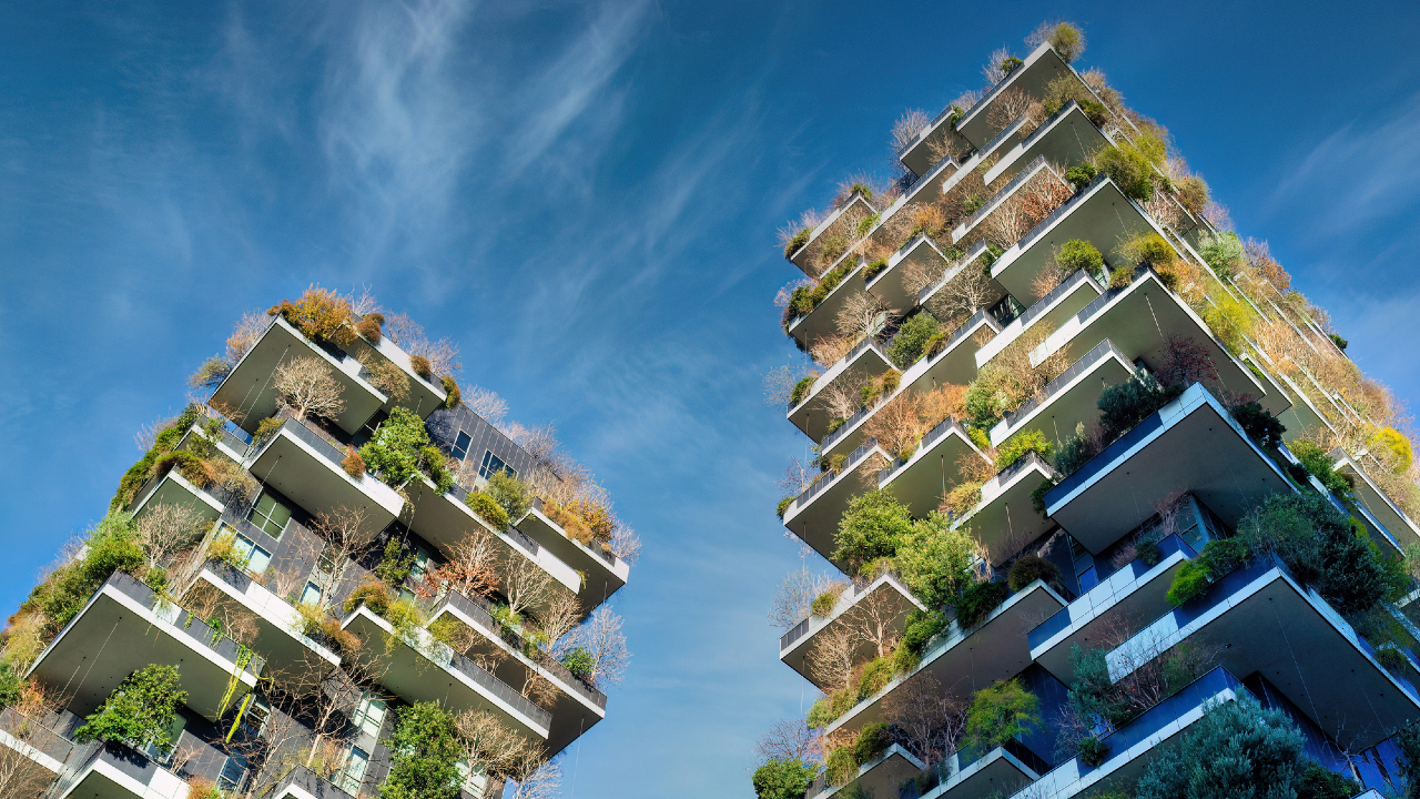 Due moderni grattacieli con vegetazione lussureggiante e alberi che crescono su più balconi, in un cielo blu brillante con nuvole ondulate.