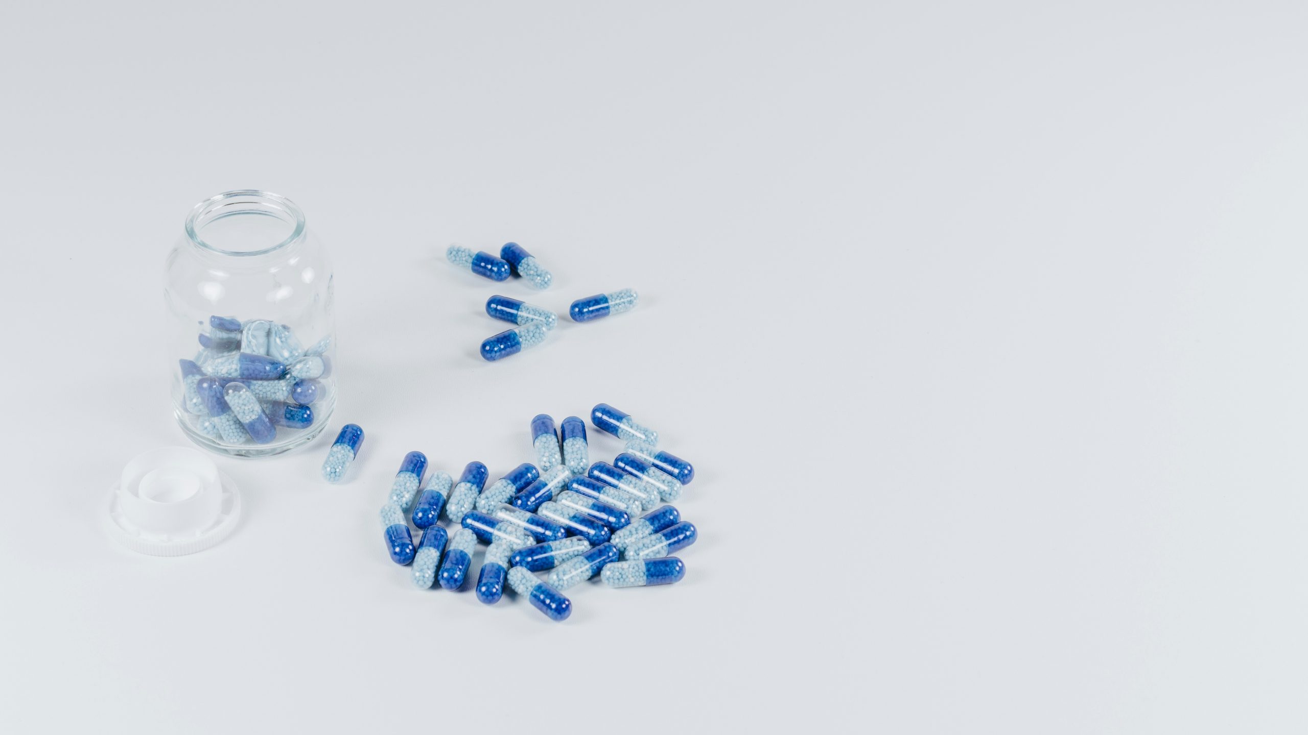 Un piccolo barattolo di vetro adagiato su un fianco, con capsule blu e trasparenti che fuoriescono su una superficie bianca. Il coperchio bianco del barattolo è posizionato vicino e diverse capsule sono sparse intorno al barattolo.