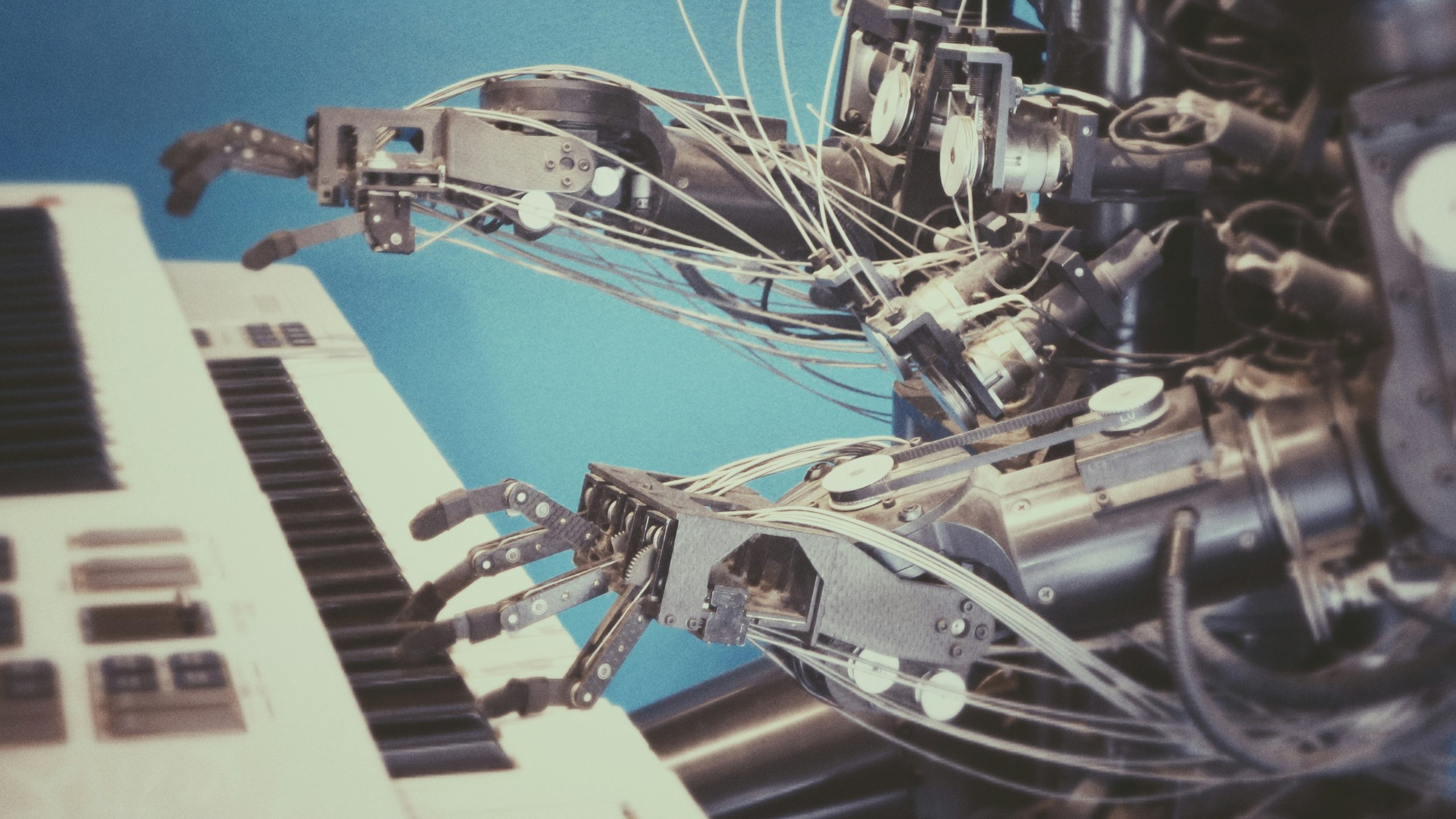 Primo piano di mani robotiche con fili e giunti metallici visibili che suonano i tasti di una tastiera elettronica su uno sfondo blu.