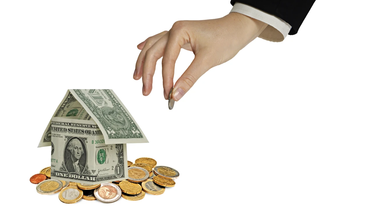 Una mano pone una moneta su un mucchio di soldi accanto a una casetta ricavata da una banconota da un dollaro Usa piegata, a simboleggiare il risparmio o l'investimento di denaro per la prima casa.