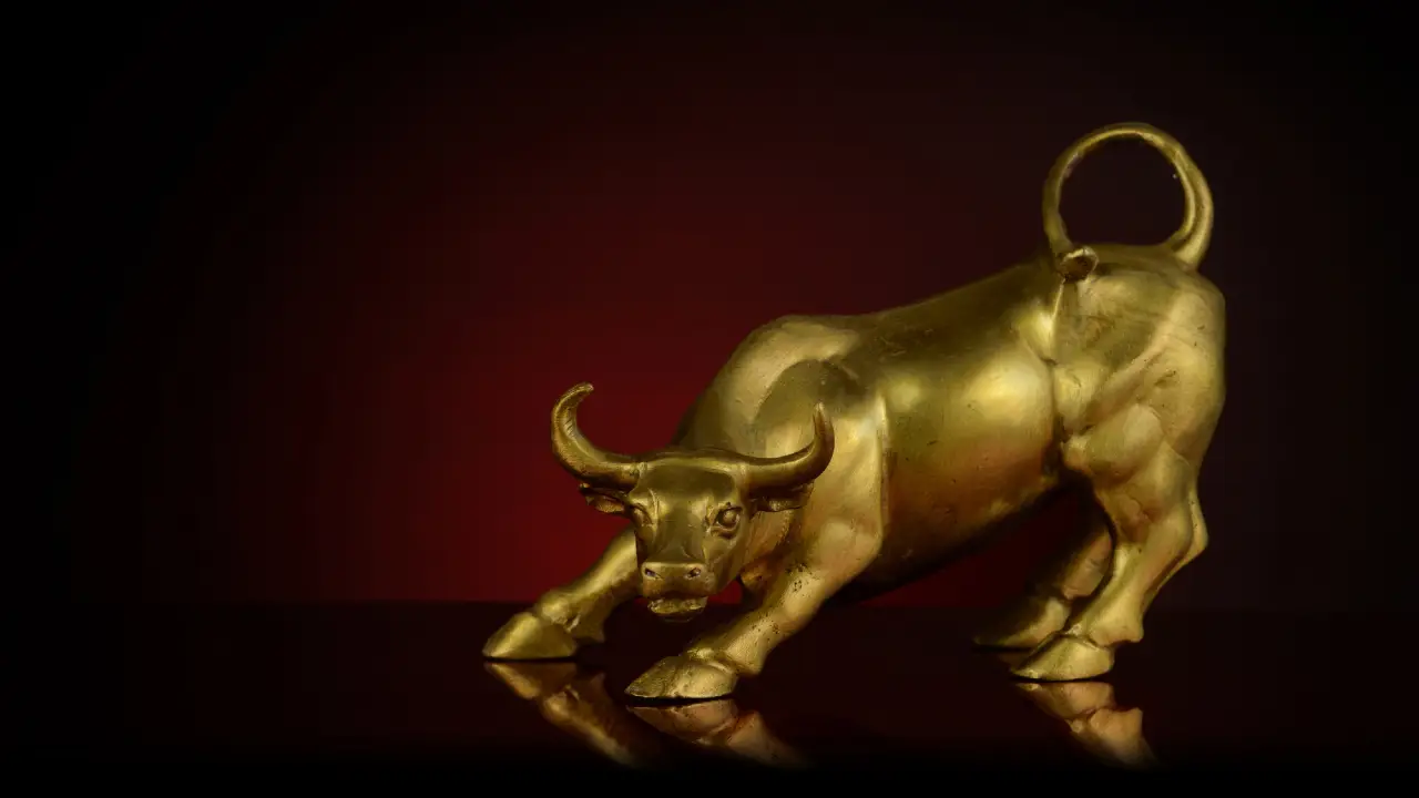 La statua di un toro dorato è posizionata in una posizione aggressiva e di carica su una superficie riflettente, con uno sfondo rosso scuro e nero sfumato.