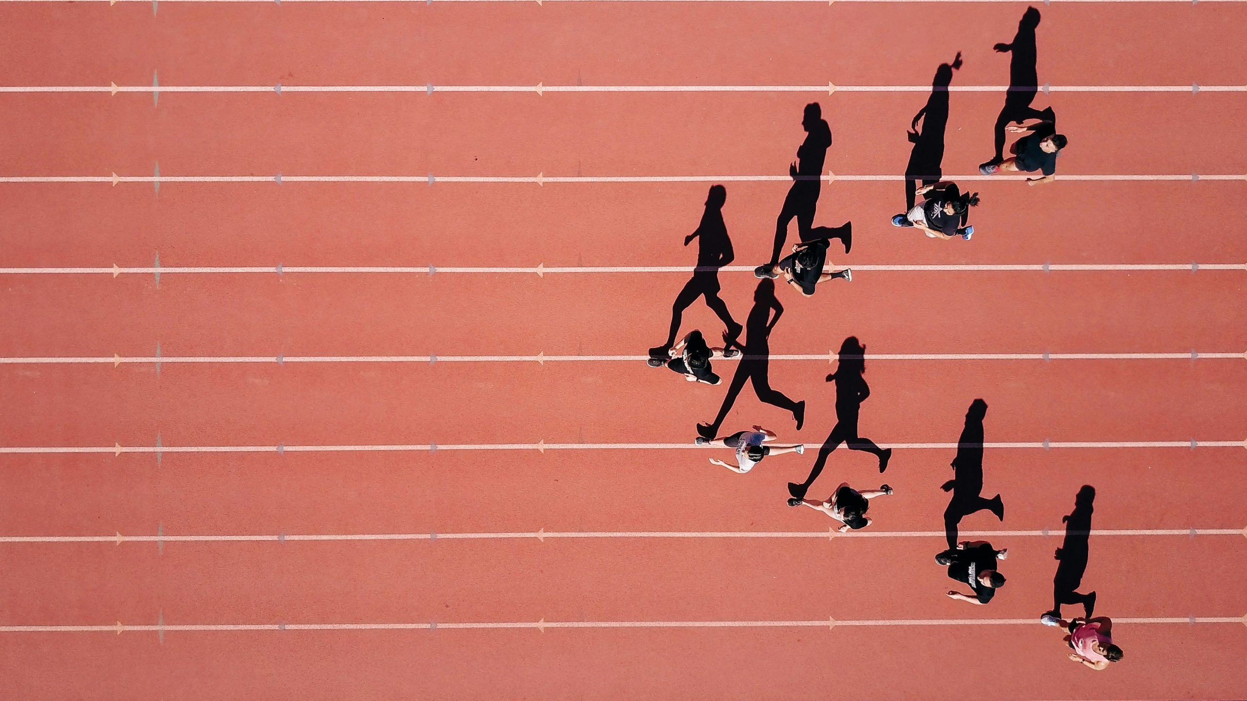 Vista aerea di diverse persone che corrono su una pista di atletica rossa, proiettando lunghe ombre sulle corsie in piena luce solare.