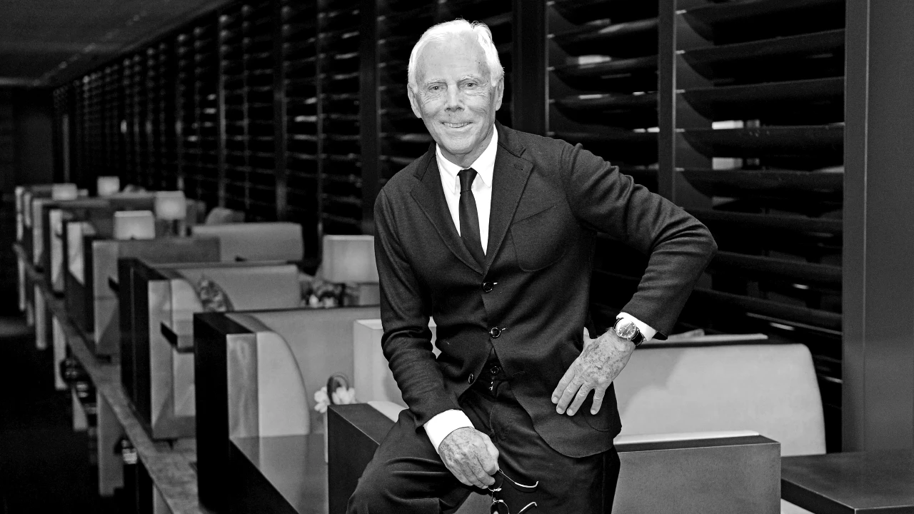 Fotografia in bianco e nero di Giorgio Armani, sorridente, in giacca e cravatta, seduto sul bordo di un tavolo in un ambiente moderno e raffinato.