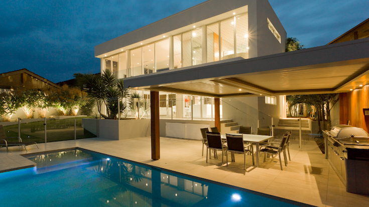 Moderna casa a due piani con ampie vetrate, illuminata al crepuscolo. Godetevi una cena all'aperto o consultate una consulenza di design nel patio con barbecue e piscina, il tutto circondato da un giardino curato e da palme.