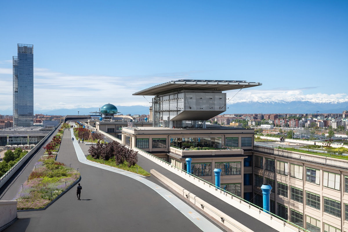 Una passerella sul tetto con verde e una moderna struttura in vetro in cima a un edificio urbano, che si affaccia su una città con montagne sullo sfondo sotto un cielo azzurro e limpido. Una persona cammina lungo il percorso.