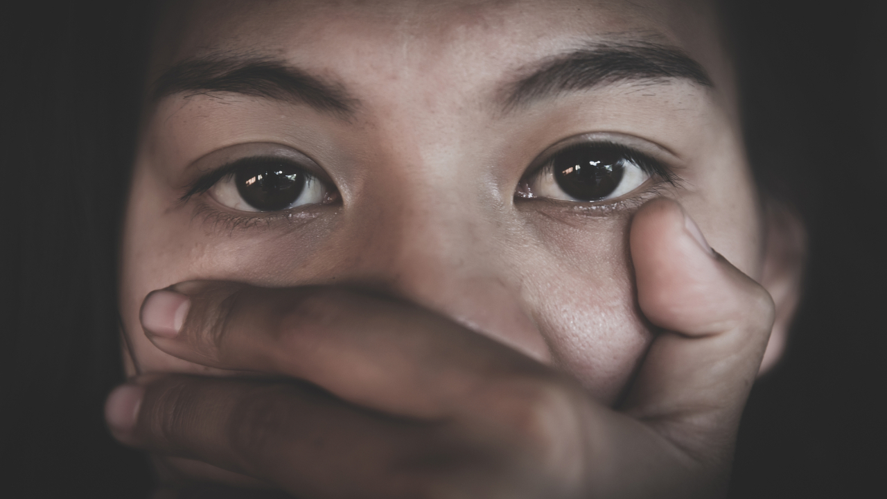 Primo piano del volto di una persona con occhi ampi ed espressivi, mentre una mano copre la bocca, trasmettendo un senso di paura, shock o silenzio. Lo sfondo è scuro e sfocato.