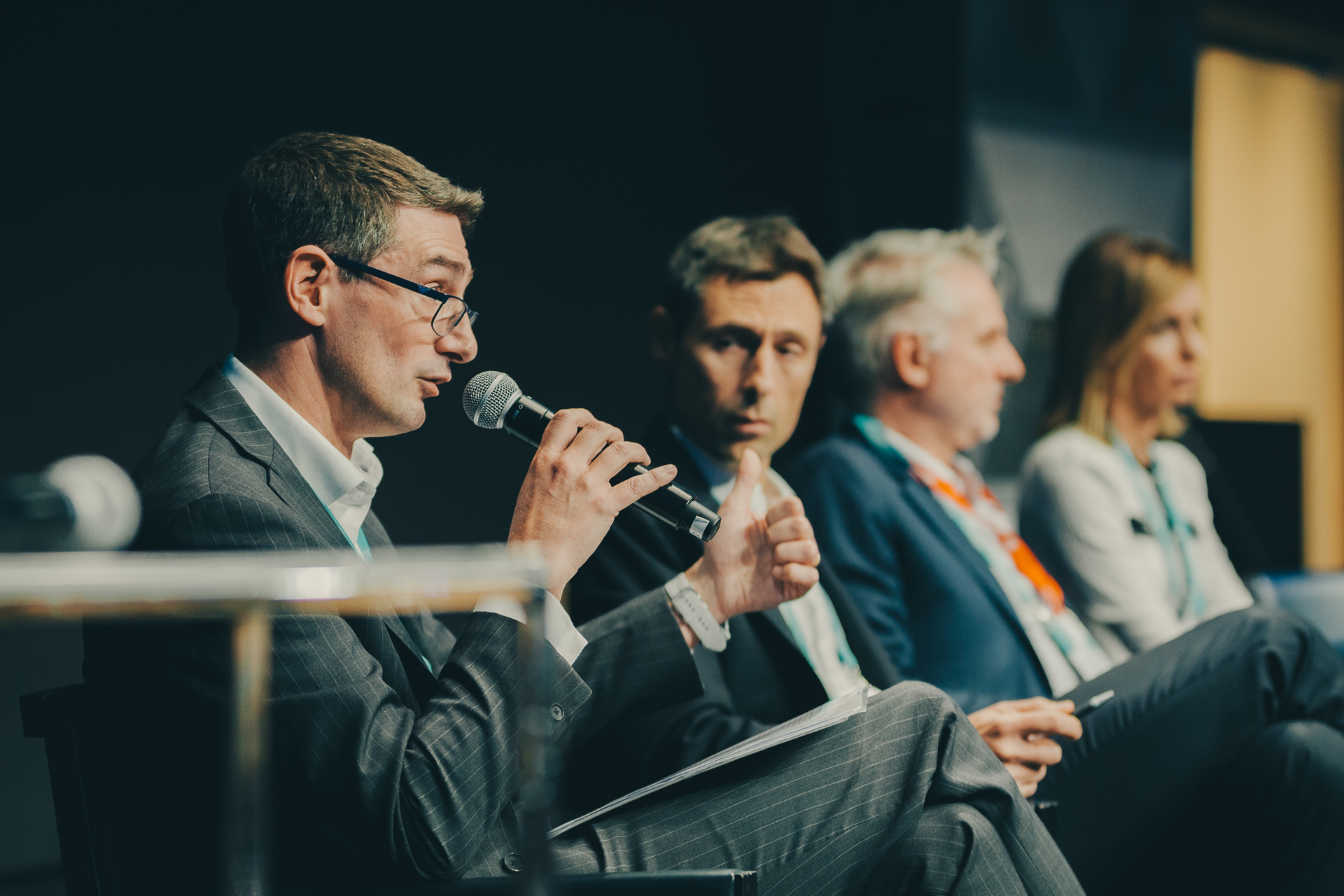 A man in a suit speaks into a microphone during a panel discussion, with three other business professionals seated beside him, all facing the audience.