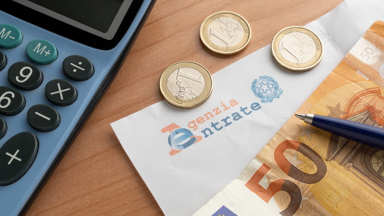 A calculator, three one-euro coins, a blue pen, a 50 euro banknote, and an envelope labeled "Agenzia Entrate" (Italian tax agency) are arranged on a wooden desk.