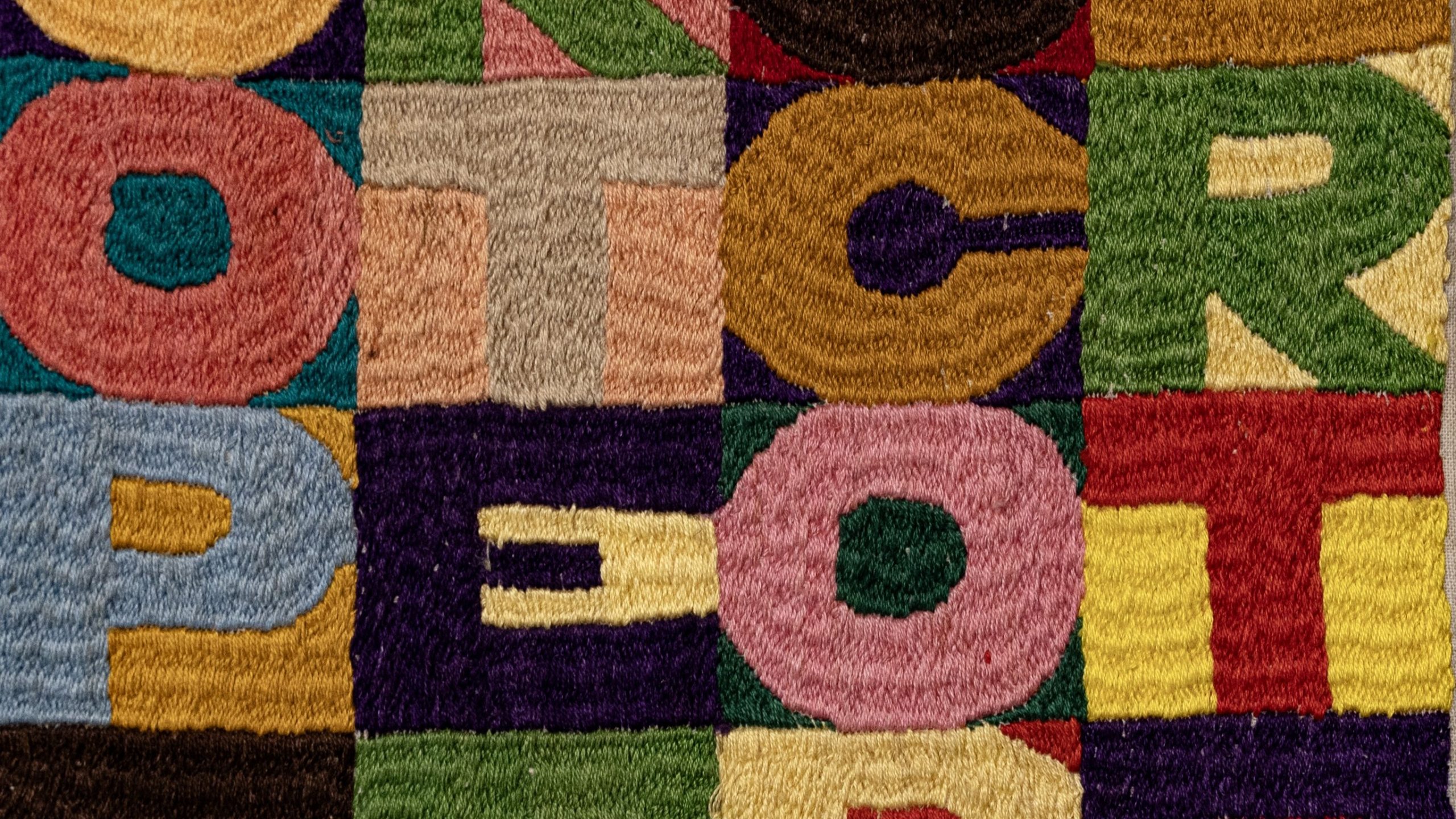 Tessuto colorato con lettere "P", "O", "T", "C" e "R" ripetute in vari colori su una griglia patchwork di sfondi contrastanti.