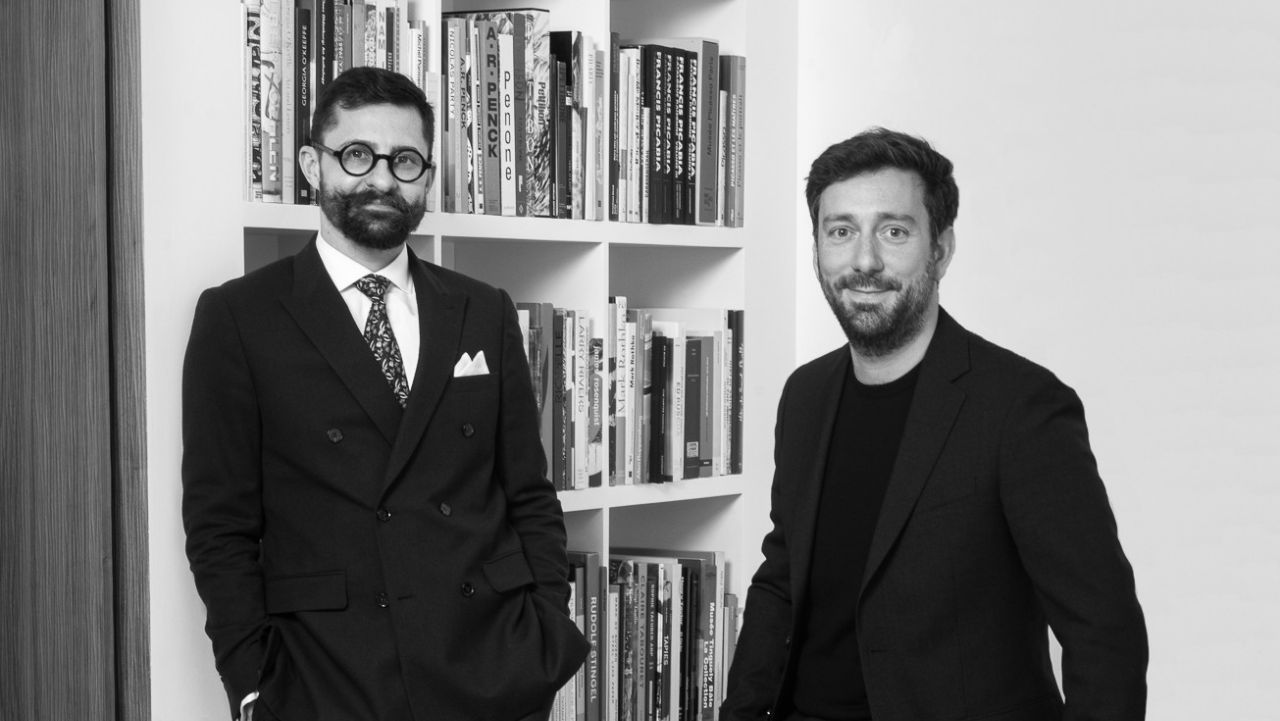 Due uomini in piedi davanti a una libreria piena di libri. Un uomo indossa giacca e cravatta e occhiali, mentre l'altro indossa giacca e camicia scura. La foto è in bianco e nero.