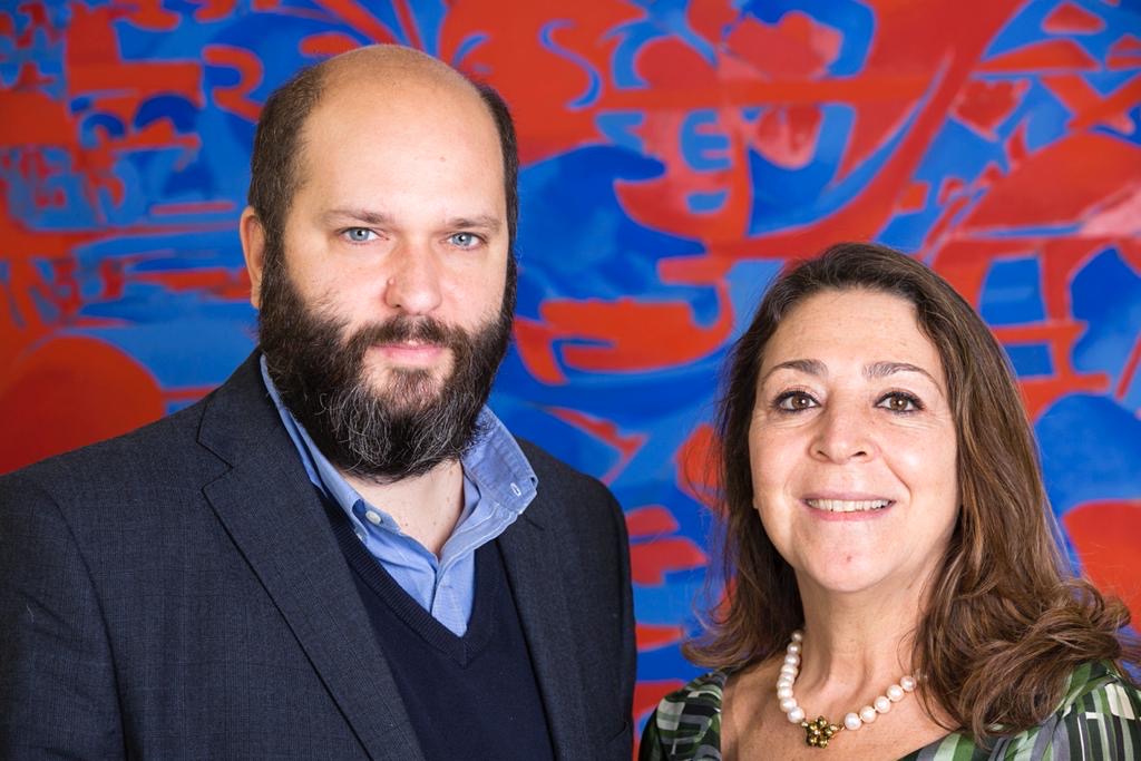 Un uomo con la barba e una donna con una collana sorridono alla telecamera, in piedi davanti a un vibrante sfondo astratto rosso e blu.