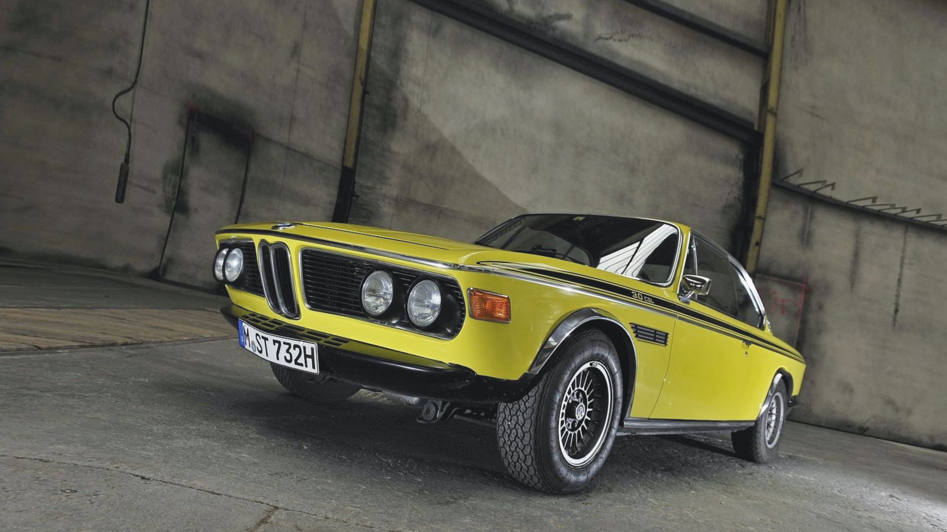 Una classica BMW 3.0 CSL coupé gialla con strisce da corsa nere è parcheggiata all'interno su un pavimento di cemento, vista da un'angolazione bassa. L'auto presenta uno stile vintage e una targa europea con la scritta "ST 732H".