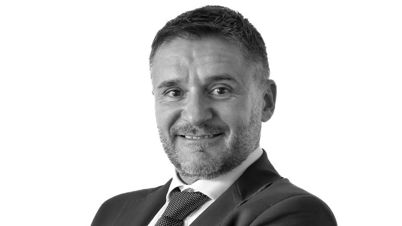Un uomo con capelli corti e barba, in giacca e cravatta, che sorride e guarda la macchina fotografica, in posa su uno sfondo bianco.
