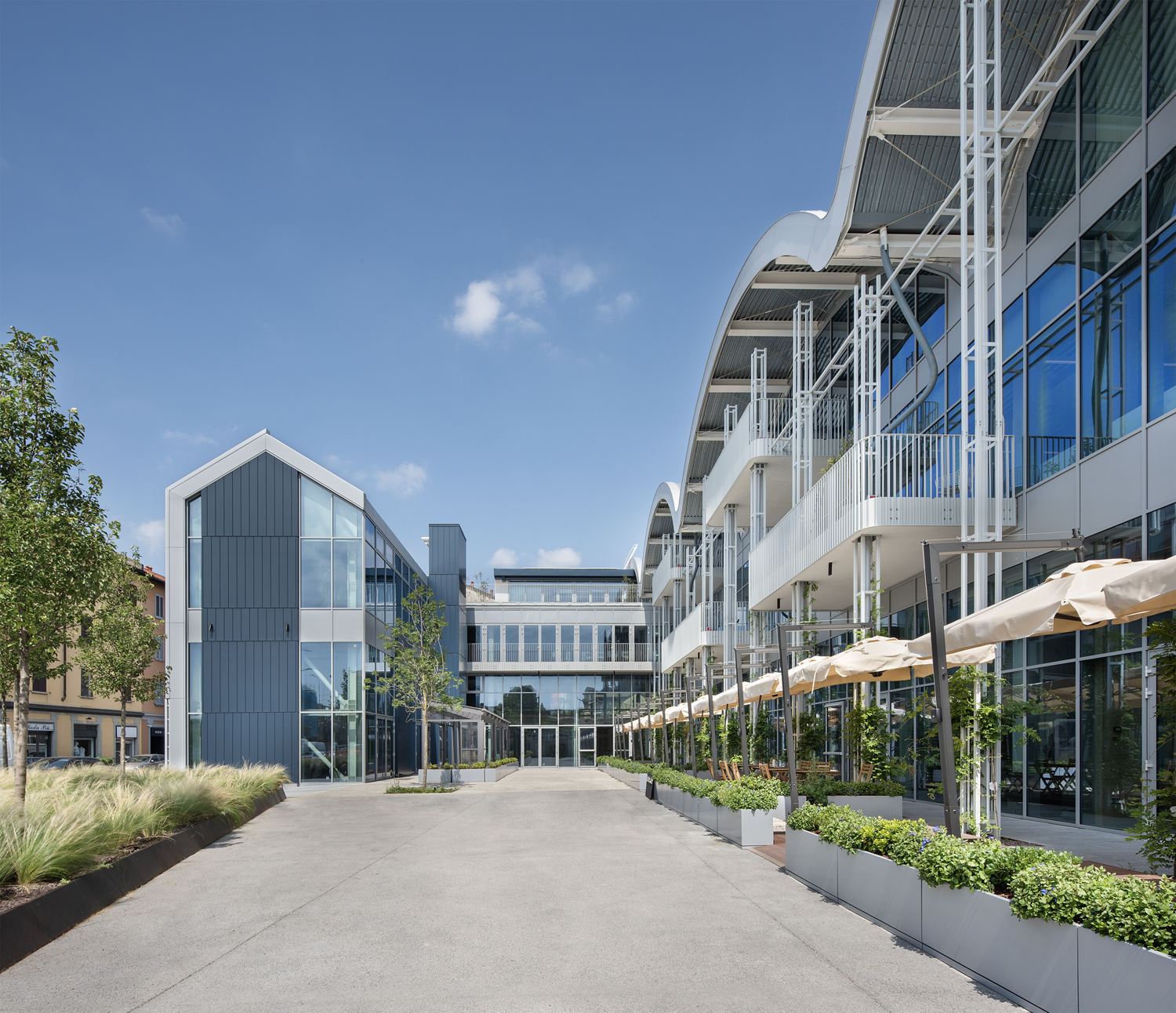 A modern commercial or office complex with curved white roofs, glass walls, outdoor seating under cream umbrellas, and landscaped greenery, all under a clear blue sky.