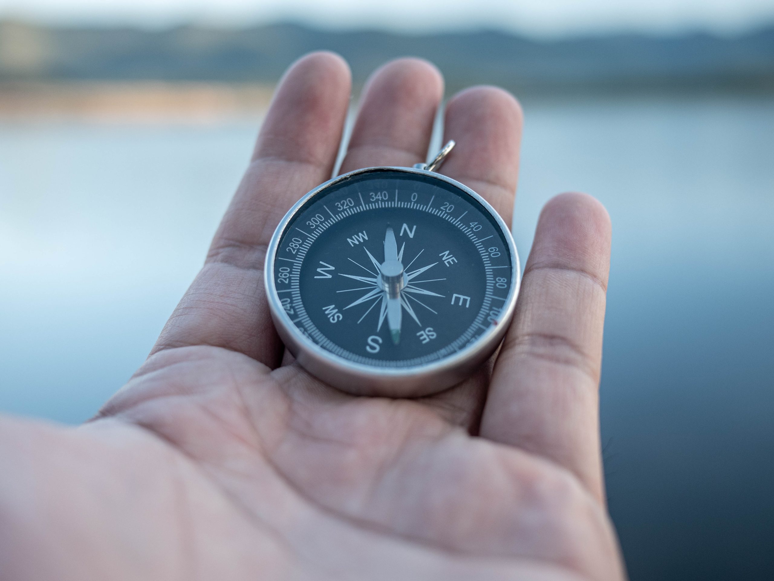 Una persona tiene una bussola d'argento nel palmo della mano, pronta a navigare su un lago calmo con montagne sfocate sullo sfondo.