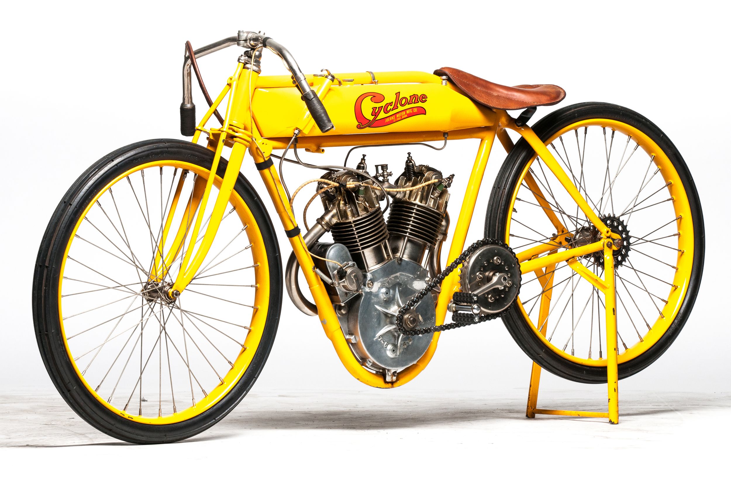 Una moto Cyclone gialla d'epoca con sella in pelle marrone e parti del motore esposte, con pneumatici sottili e parafanghi minimi, esposta su un cavalletto su sfondo bianco.