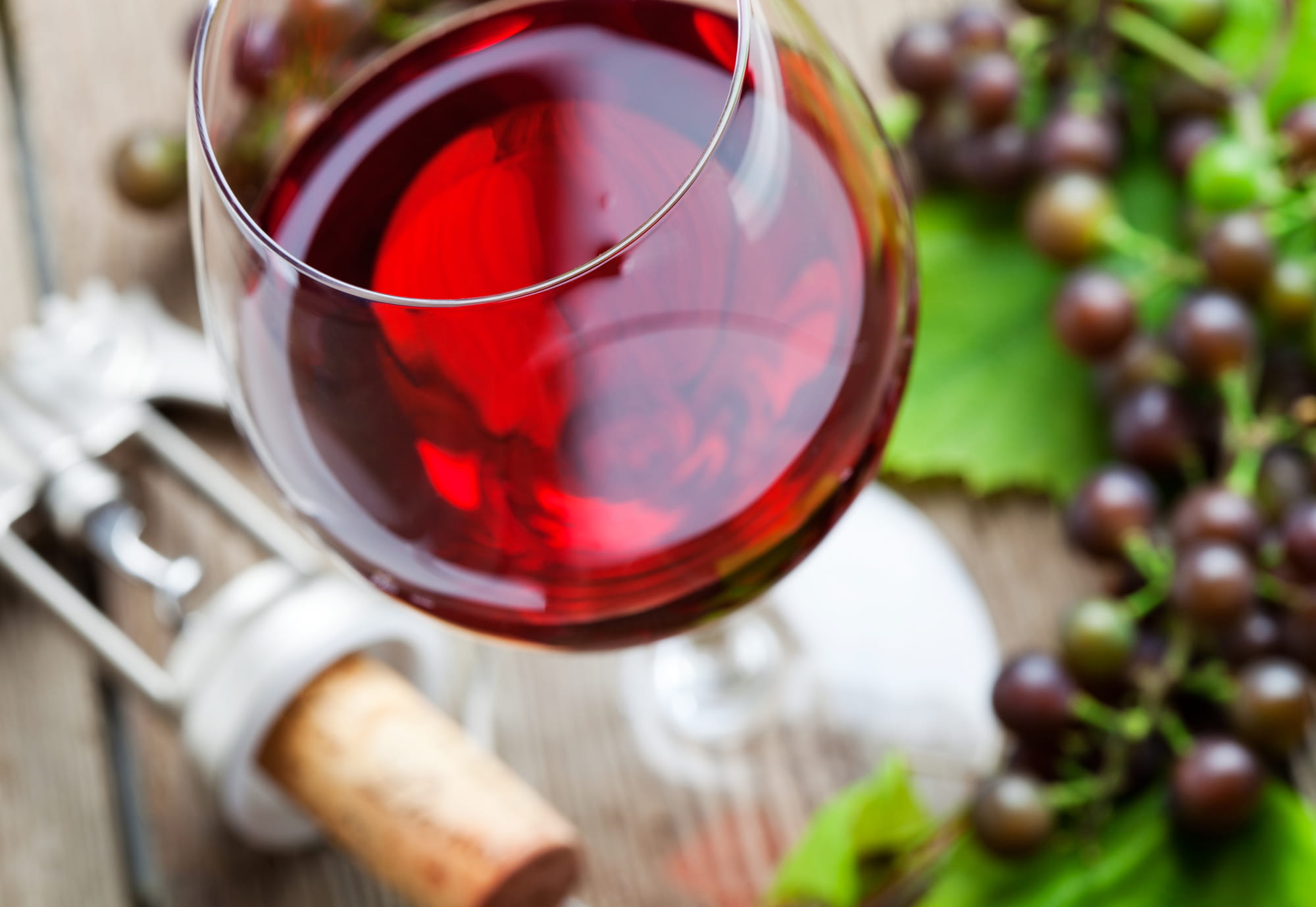 A close-up of a glass of red wine next to a corkscrew, a cork, and a bunch of grapes with green leaves on a wooden surface.