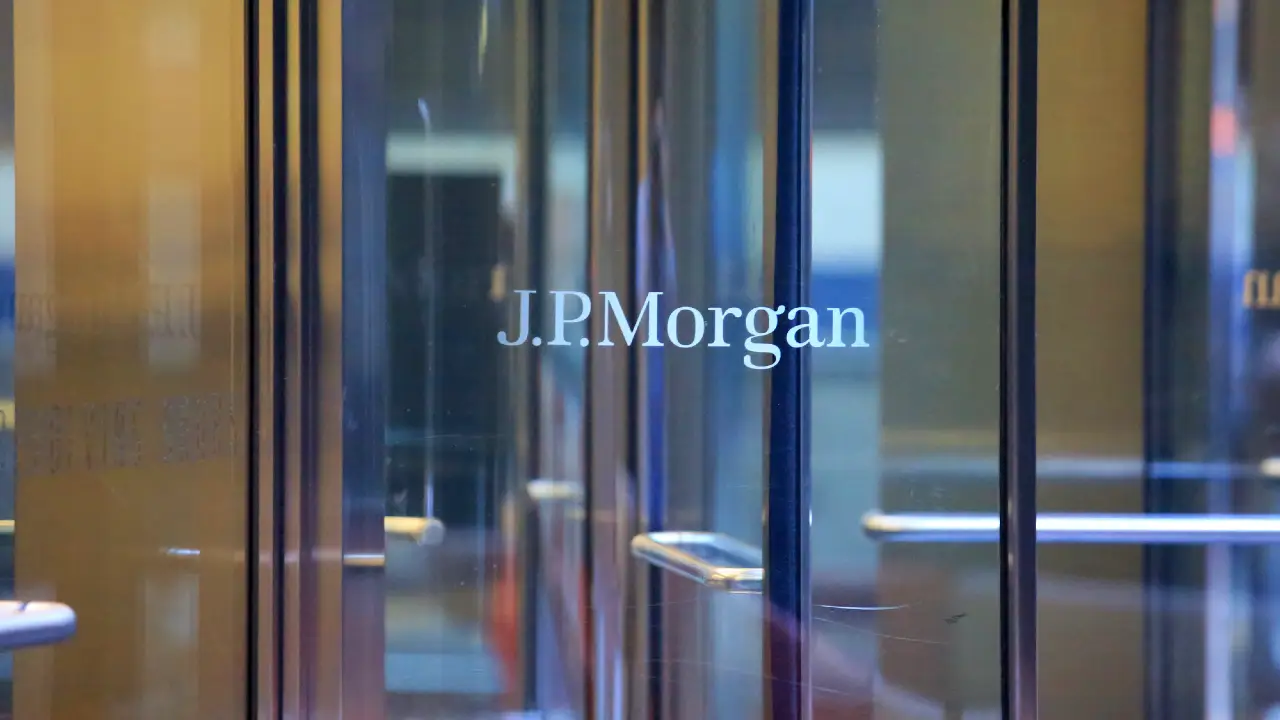 Porte di vetro con il logo "J.P. Morgan" in lettere bianche, che riflettono la luce e gli edifici circostanti, probabilmente all'ingresso di un ufficio aziendale.