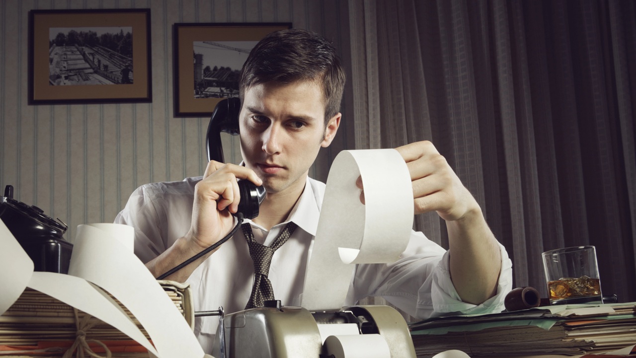 Un uomo in camicia e cravatta siede a una scrivania disordinata, tenendo in mano un telefono ed esaminando una lunga ricevuta, forse rivedendo documenti fiscali come "In arrivo una nuova rottamazione (quinques) per definire i debiti con il fisco.