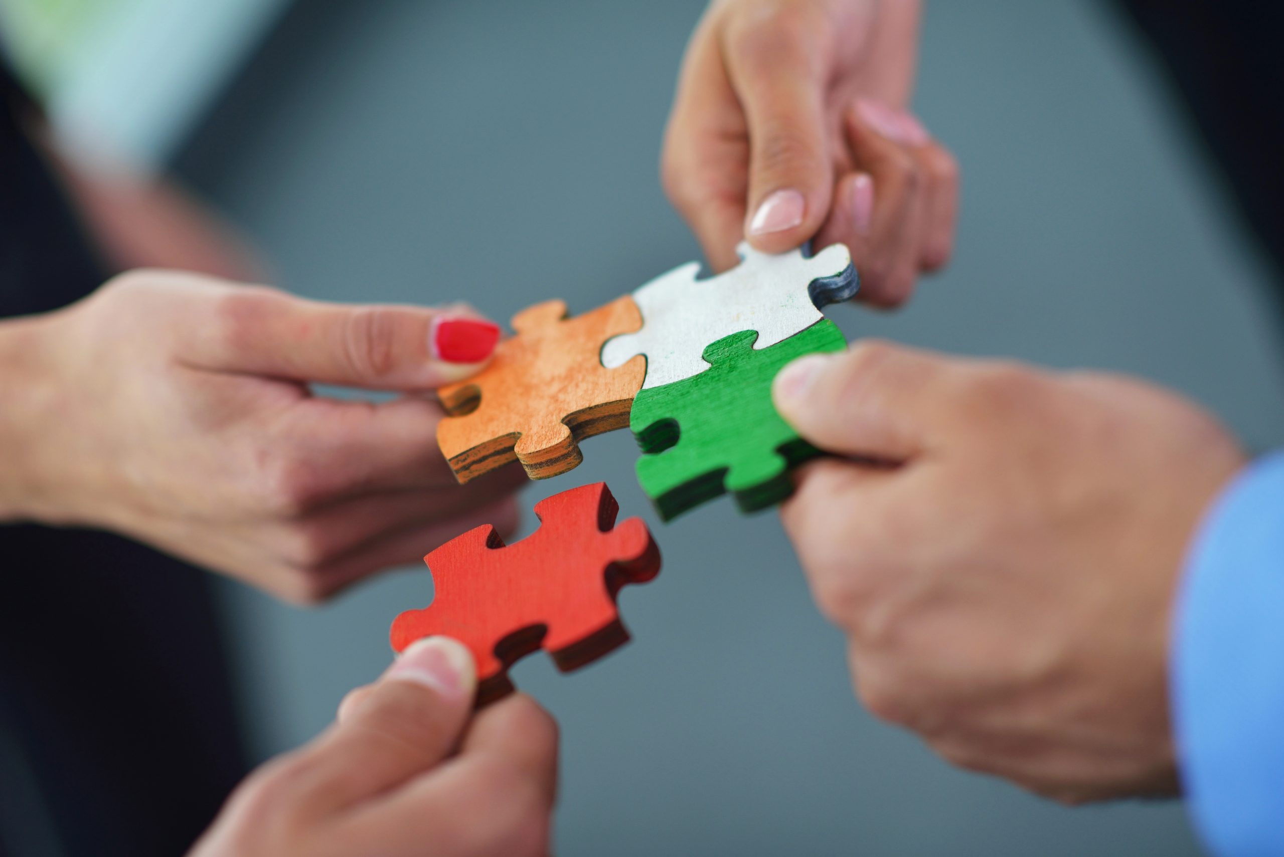 Quattro persone tengono ciascuna un pezzo di puzzle di colore diverso - arancione, bianco, verde e rosso - e lavorano insieme per collegare i pezzi, simboleggiando il lavoro di squadra e la collaborazione.