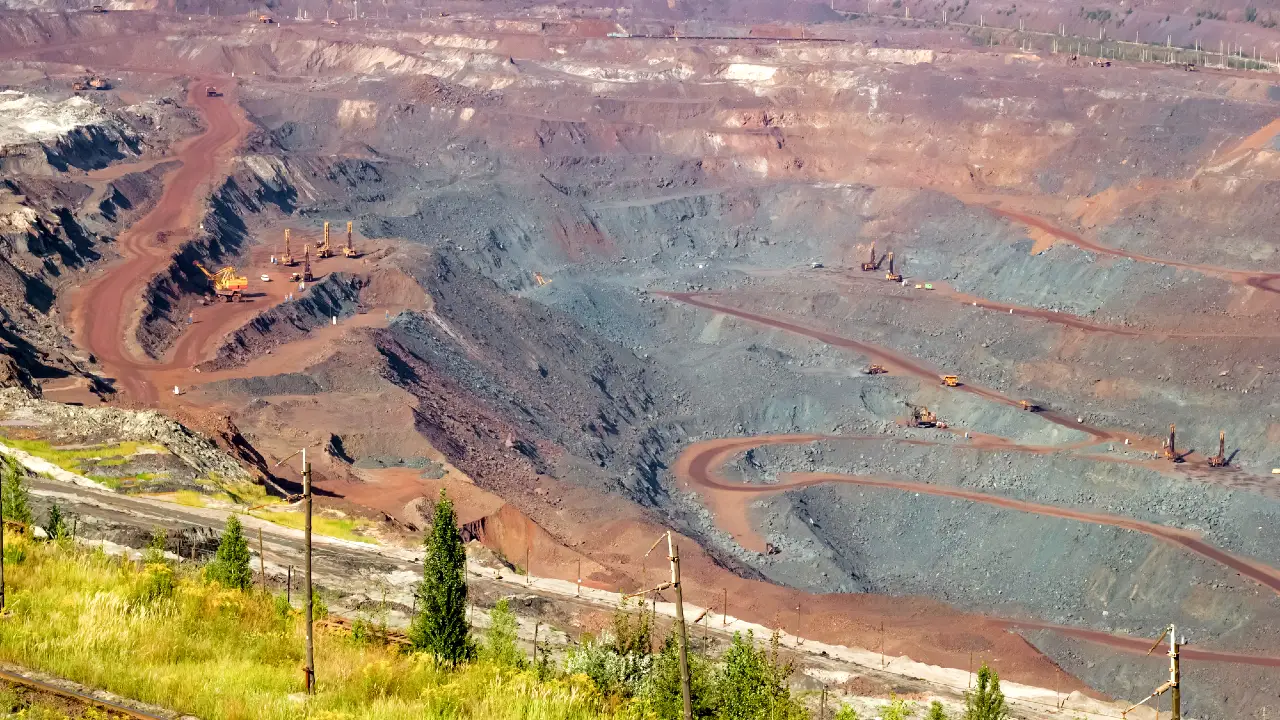 Un'ampia miniera a cielo aperto con pendii terrazzati e strade sterrate tortuose. Sono visibili diversi grandi veicoli e macchinari minerari, circondati da macchie di vegetazione in primo piano.