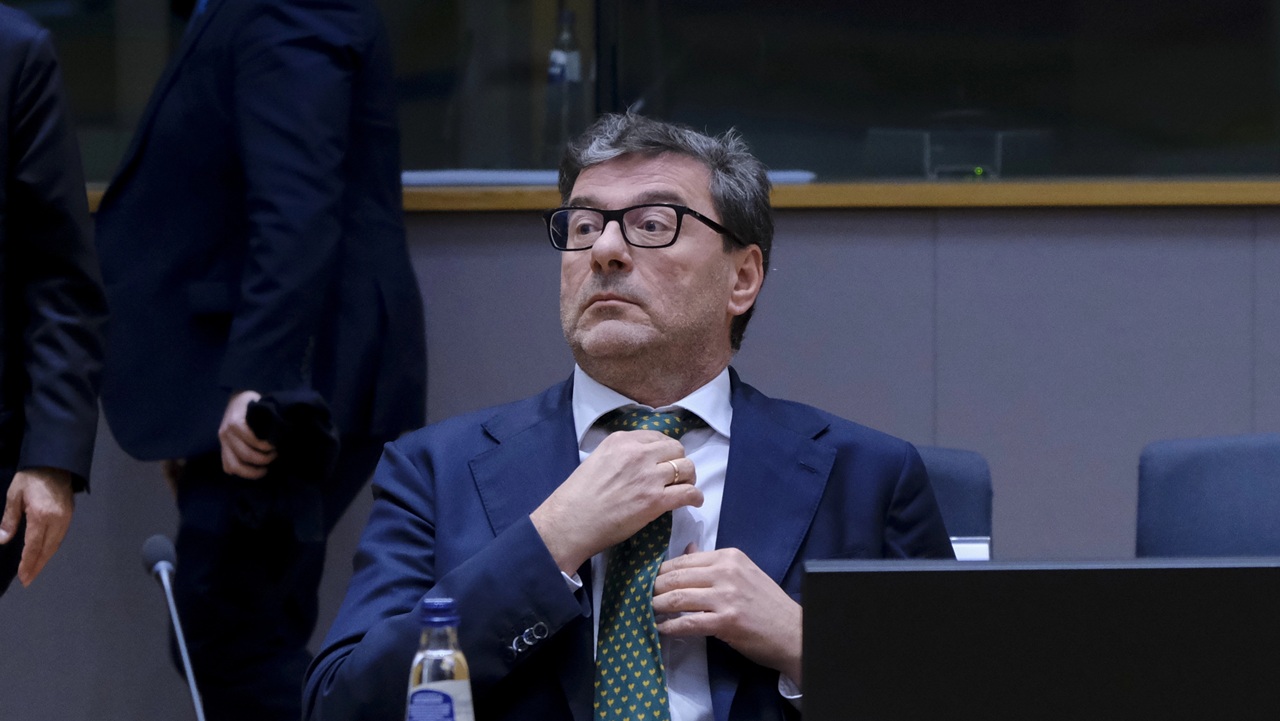 Un uomo in abito e occhiali si aggiusta la cravatta verde a pois seduto a un tavolo da conferenza. Davanti a lui ci sono una bottiglia d'acqua e il monitor di un computer, mentre altre persone sono sfocate sullo sfondo.