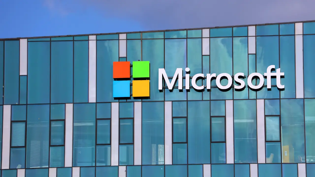 L'esterno di un edificio moderno con un grande logo Microsoft e un'icona colorata sulle vetrate, sotto un cielo blu.